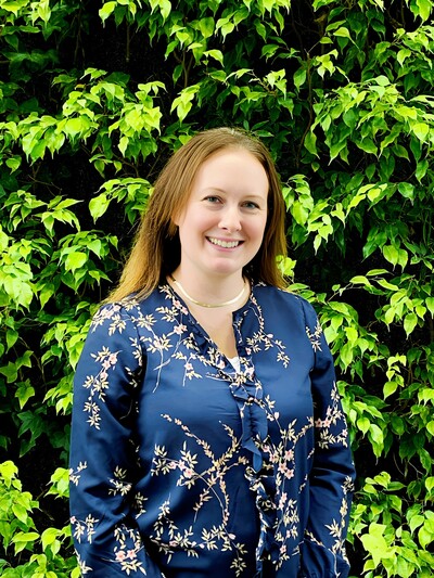 Profile picture of jenny standing in front of bright green leaves covered on a wall and she is smiling.
